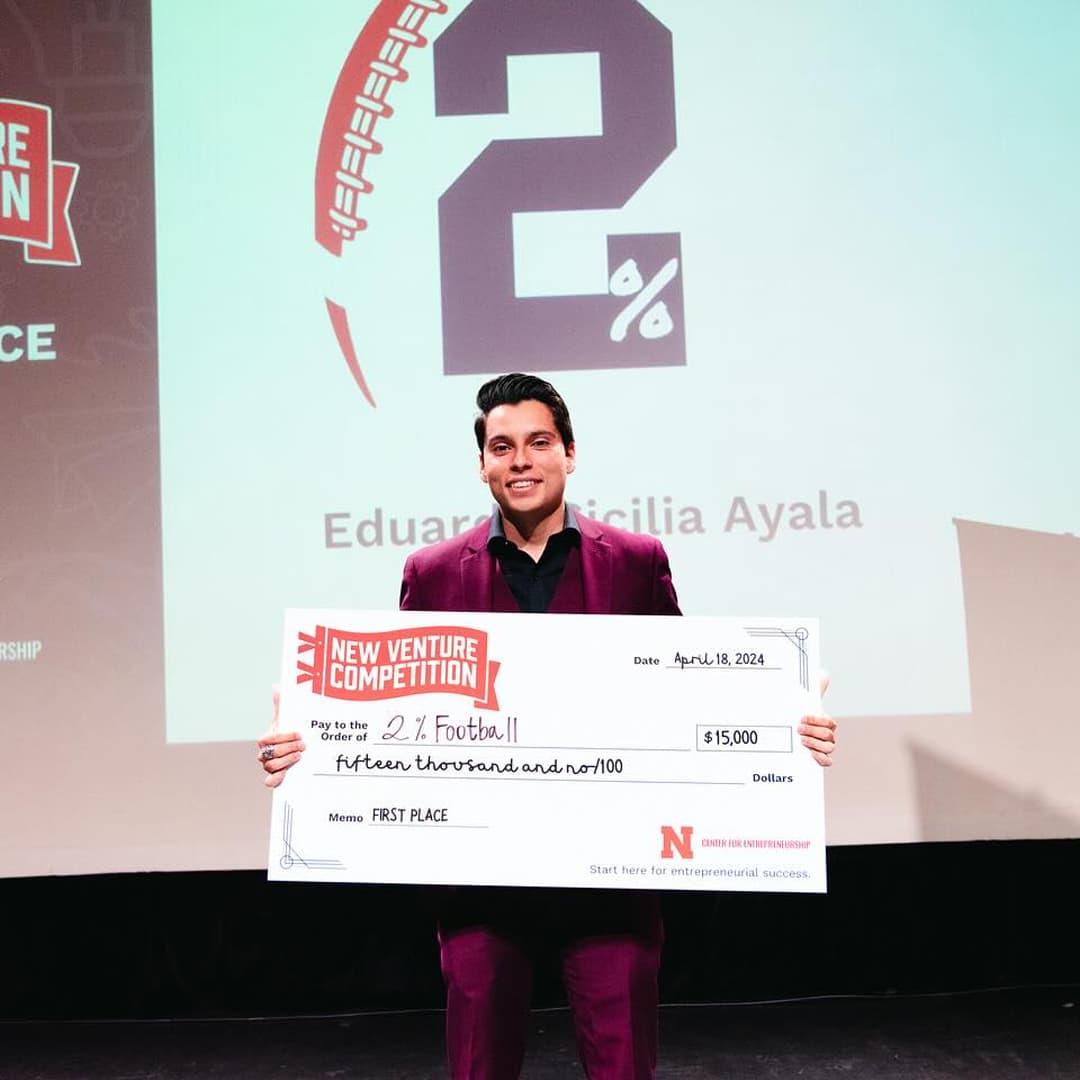 Husker holds an oversized $15,000 check from New Venture Competition payable to 2% Football.