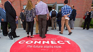 A group of people enter Nebraska Innovation Campus for a tour of the facilities. A large circle decal on the floor reads, 'Innovation lives here. Connect. The future starts now.'