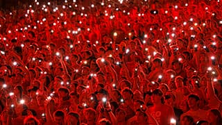 At a nighttime Nebraska football game, the crowd, some holding up phone flashlights, is bathed in red light from above.