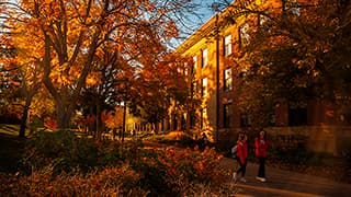 Two students wearing backpacks talk while walking across the tree-lined campus among on a sunny fall day.