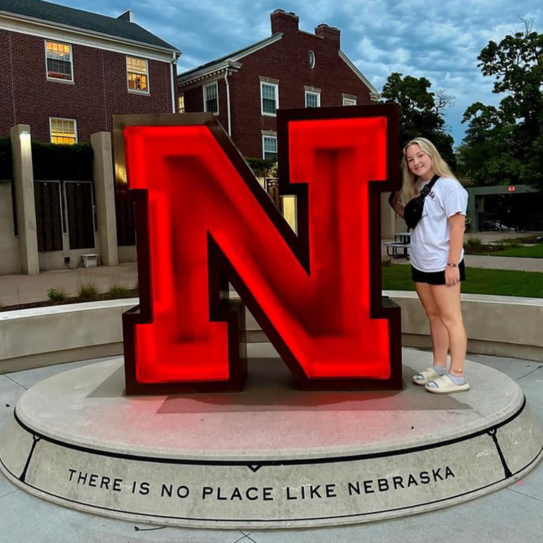 Smiling Husker poses next to red, illuminated N. Below the N reads There is No Place Like Nebraska .