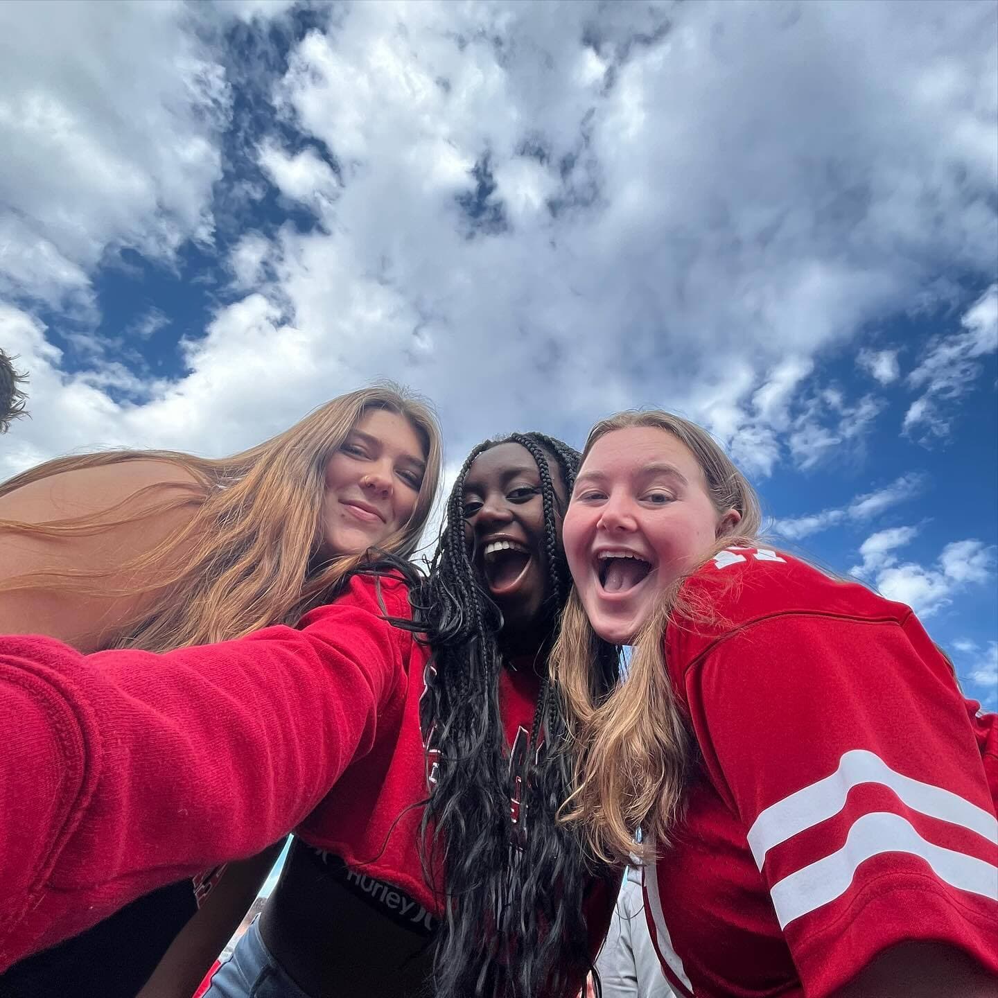 Three smiling Huskers take a selfie with cloudy skies in the background.