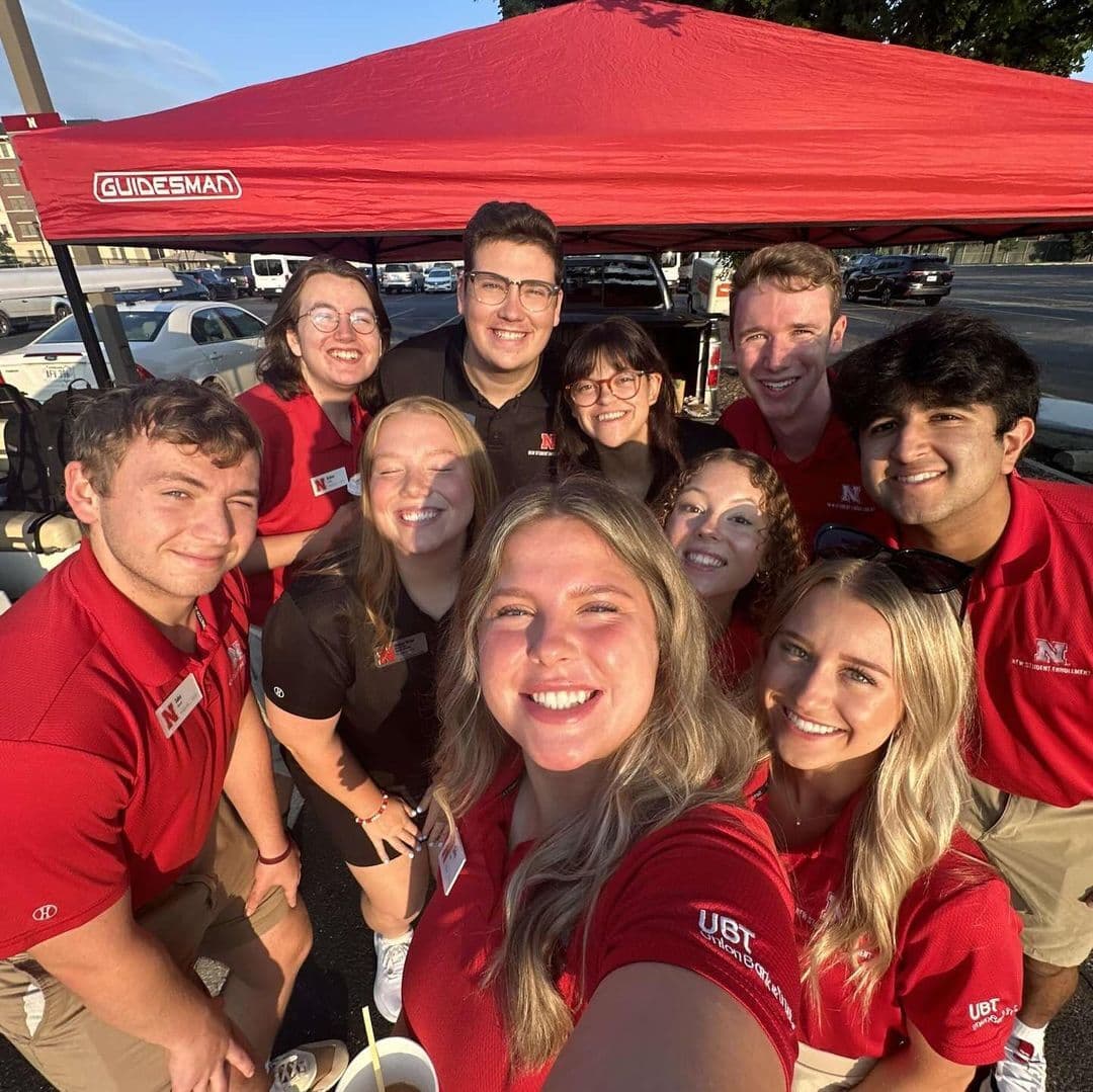 A group of ten smiling Huskers take a selfie in front of a tent in a parking lot.