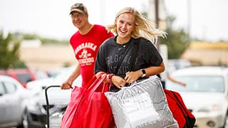 A smiling, new student carries an arm-load of belongings while her father pulls a cart behind them.