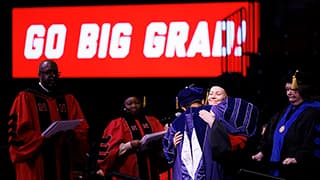 At Winter Commencement, a Husker graduate hugs someone onstage while receiving their degree. An illuminated red-and-white digital sign that reads Go Big Grad shines in the background.