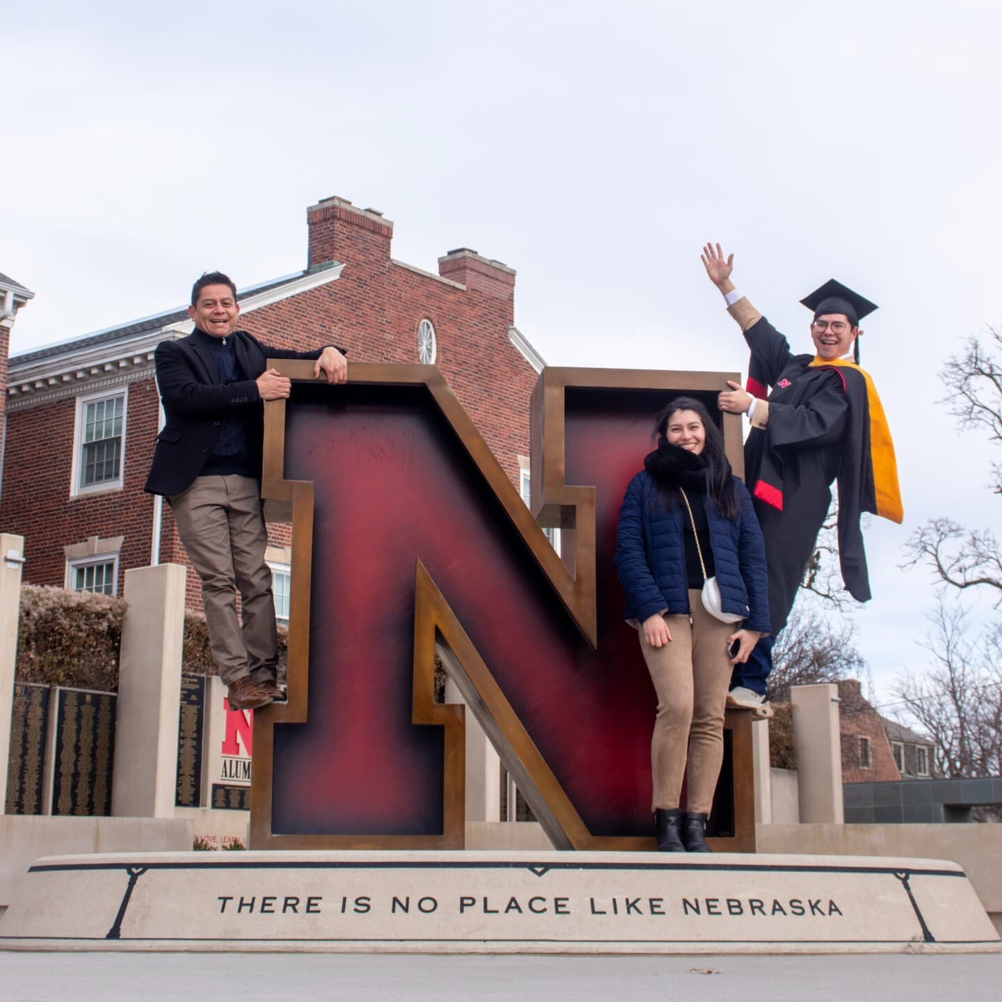 Three people, including a Husker grad in cap and gown, pose on the Nebraska N in front of Wick Alumni Center.