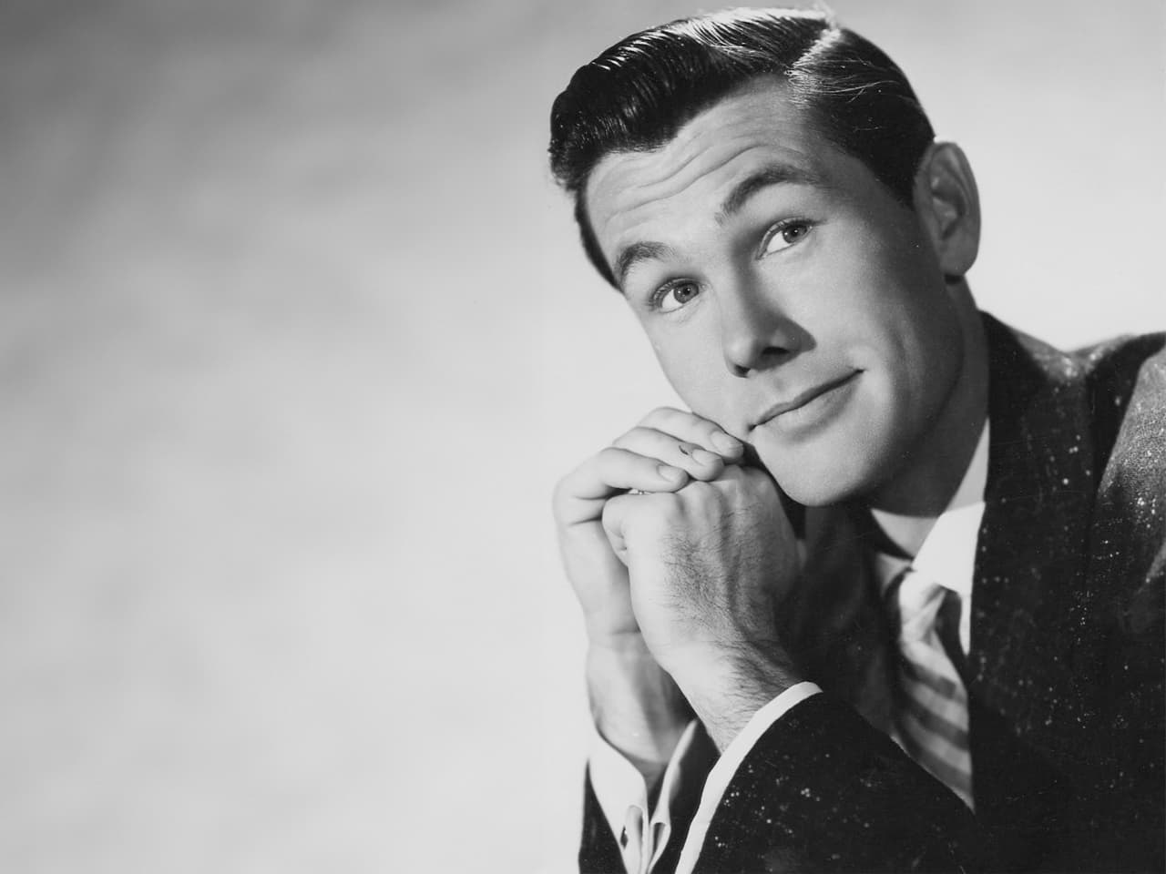 1957 black and white publicity portrait of Johnny Carson with his hands clasped near his face with a slight smile and looking off into the distance