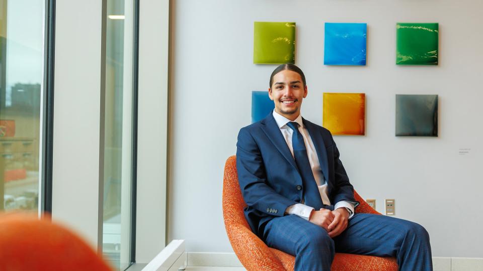 Bennett Yellow Bird III, wearing sport coat and tie, sits in an orange chair in front of a grid of six small mostly solid color paintings arranged in a grid